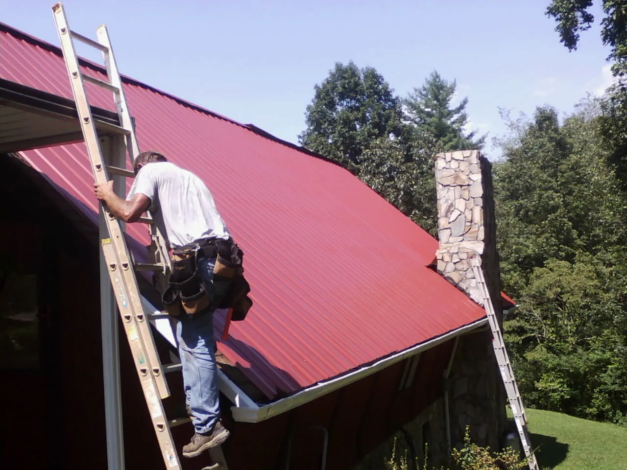 Roofer on a ladder beside a red metal roof and stone chimney — Soffit & Fascia Repair by Roofing Brothers in Rosalia